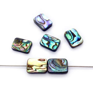 May include: Rectangular abalone shell beads in iridescent blues, greens, and purples. The beads are strung on a thin wire, with some scattered around. The beads have a polished, glossy finish, and are set against a white background.