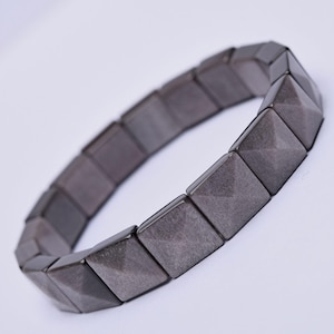 May include: A dark grey bracelet with square, pyramid-shaped beads. The bracelet is made of a metallic material and is designed to be worn around the wrist. The beads are connected by an elastic band.