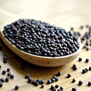 May include: A wooden spoon filled with black mustard seeds. The seeds are scattered on a wooden surface.