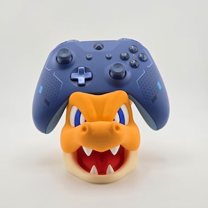 May include: A blue video game controller rests on top of an orange and cream-colored character-themed controller stand. The stand features a detailed, cartoonish design with sharp teeth and blue eyes. The controller has a cross-shaped directional pad.