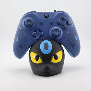 May include: A blue Xbox controller rests on a black and yellow Umbreon-themed controller stand. The controller has a cross-shaped directional pad and various buttons. The stand features yellow eyes and a blue ring.