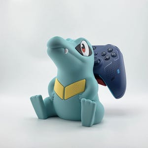 May include: A teal-colored figurine of a cartoon crocodile with a yellow chest design, holding a blue video game controller. The controller is dark blue with black buttons. The figurine is sitting on a white surface.