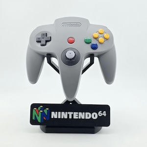 May include: A grey Nintendo 64 video game controller with a dark grey directional pad, a central white analog stick, and four colored action buttons in yellow, green, and blue, mounted on a black display stand featuring the Nintendo 64 logo.