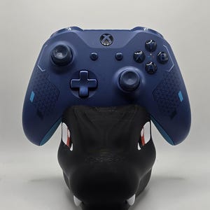 May include: A blue Xbox controller with a geometric pattern on the back, resting on a black stand with red accents.