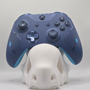May include: A navy blue Xbox controller rests on a white, stylized skull-shaped stand. The controller has a blue cross-shaped directional pad and black control sticks. The stand is a solid, smooth, and matte white.