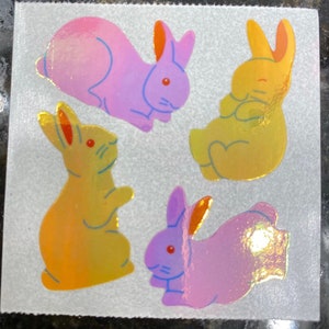 May include: A sheet of four holographic bunny stickers in shades of pink, yellow, and orange. Each bunny has a different pose, with blue outlines and red eyes. The stickers are on a white background.