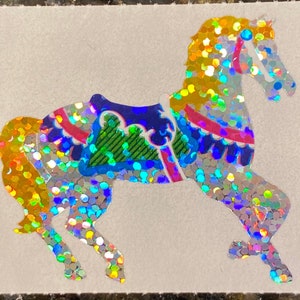 May include: A sticker of a carousel horse with a shimmering, glittery design. The horse features a saddle with blue, pink, and green accents. The horse's body is covered in iridescent glitter, reflecting a rainbow of colors.