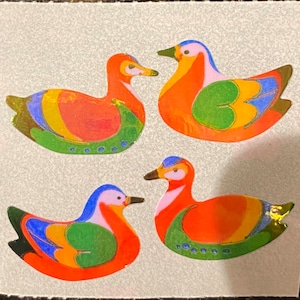 May include: Four colorful duck stickers. Each duck has a vibrant orange body, with accents of blue, green, yellow, and pink. The stickers have a gold foil finish on the wings and tail feathers. The ducks are arranged on a light gray background.