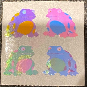 May include: Four colorful frog stickers with iridescent, holographic effects. Each frog has a unique color scheme: purple with yellow spots, pink with blue spots, green with pink spots, and blue with purple spots. The stickers are on a square, off-white background.