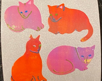 May include: Four colorful cat stickers. Two are pink, one is orange, and one is a gradient of orange and pink. Each cat has a simple, smiling face with blue eyes and gold accents. The stickers are on a light gray background.