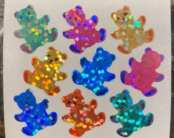May include: A sheet of holographic stickers featuring colorful teddy bears. The bears are in various colors, including blue, pink, gold, and green, with a shiny, iridescent finish. Each bear has a smiling face and is decorated with small, colorful dots.