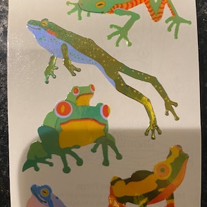 May include: Sheet of colorful frog stickers. The stickers feature various frog designs in shades of green, yellow, orange, and blue. Some frogs have iridescent details. The stickers are on a white background.