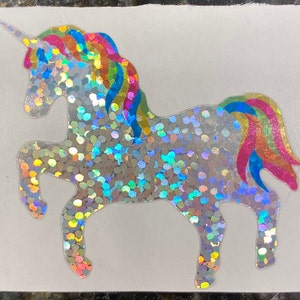 May include: A holographic unicorn sticker with a rainbow mane and tail. The unicorn is silver with iridescent glitter accents. The mane and tail feature a vibrant spectrum of colors, including pink, yellow, blue, and green.