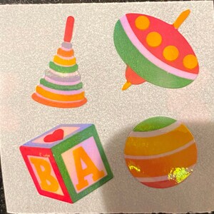 May include: A collection of colorful toy stickers. Includes a stackable ring toy, a spinning top, a cube block with the letters B and A, and a striped ball. The stickers are in shades of red, orange, yellow, green, and purple.
