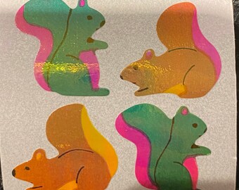 May include: Four holographic squirrel stickers. Each squirrel has a different color scheme, with green, orange, and gold bodies and pink and yellow accents on their tails. The stickers are on a white background.
