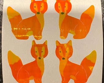 May include: Four orange fox stickers with yellow accents. Each sticker features a stylized fox with a pointed snout, upright ears, and a bushy tail. The stickers are on a white background.