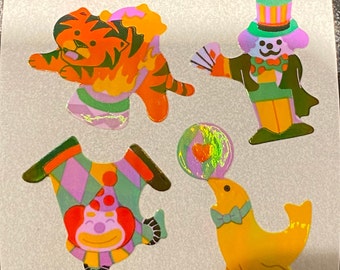 May include: A sheet of colorful circus-themed stickers. The stickers include a tiger, a dog in a top hat, a clown, and a seal balancing a ball. The stickers have a holographic, shiny finish.