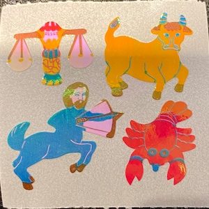 May include: Four holographic stickers featuring zodiac symbols: Libra scales, a golden bull, a blue Sagittarius centaur with a bow and arrow, and a red crab. The stickers have a shiny, iridescent finish and are set against a light gray background.