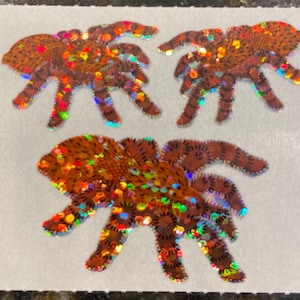 May include: Three orange and brown spider-shaped stickers with iridescent glitter accents. The stickers have detailed leg and body patterns, and the glitter reflects multiple colors. The stickers are on a white background.