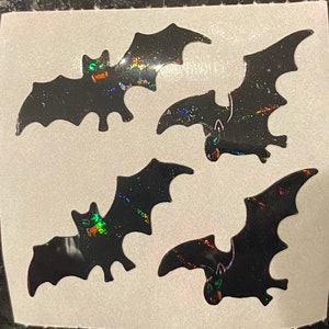 May include: Five black bat stickers with iridescent accents. The bats have detailed wing shapes and are arranged on a white background. These stickers are suitable for Halloween decorations or craft projects.