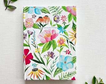 May include: A white notebook with a floral design featuring pink, blue, yellow, and red flowers. The text "Hannah Mary Design" is printed on the front cover.