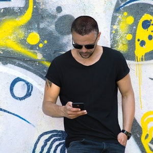 Puede incluir: Un hombre con gafas de sol y una camiseta negra se encuentra frente a una pared con grafitis, mirando un teléfono inteligente en su mano.