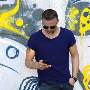 Puede incluir: Un hombre con gafas de sol y una camiseta azul se encuentra frente a una pared con grafitis, mirando un teléfono inteligente en su mano.
