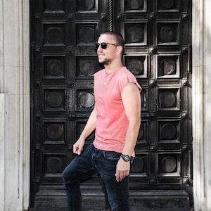 Puede incluir: Un hombre con una camiseta rosa, pantalones vaqueros azules oscuros y gafas de sol se encuentra frente a una gran puerta de madera.