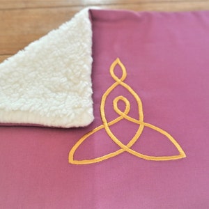Puede incluir: Una manta suave de color rosa con un diseño de nudo celta bordado en oro. La manta tiene un forro de sherpa blanco.