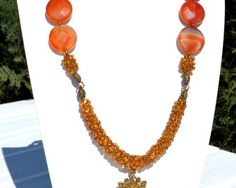 May include: Orange and gold beaded necklace with large, round, faceted orange beads. The necklace features a gold filigree pendant with a small orange bead drop. The necklace is displayed on a white bust.