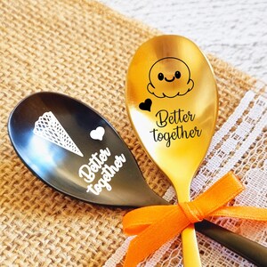 Può includere: Due cucchiai dorati e neri con le parole "Better Together" incise su di essi. I cucchiai sono su uno sfondo di juta con un rivestimento in pizzo bianco. Un cucchiaio ha un piccolo disegno di cono gelato e l'altro ha un cuore.