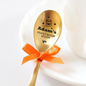Könnte beinhalten: Ein goldfarbener Löffel mit einer schwarzen Aufschrift "Adam's Peanut Butter Spoon" und einem kleinen Peanut-Gläser-Symbol. Der Löffel ist mit einem orangefarbenen Band gebunden.