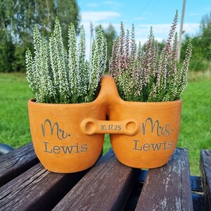 Pode incluir: Um vaso de terracota com dois vasos conectados, cada um com plantas floridas. Um vaso está inscrito com "Mr. Lewis", o outro com "Mrs. Lewis", e a data "10.17.25" está entre eles. As plantas têm flores brancas e rosas.