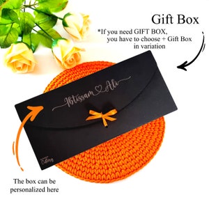 May include: A black gift box with a white ribbon and the text "Ibtissam Ali" on the front. The box is sitting on a bright orange crocheted mat. The text "Gift Box" is written in white on a black background above the box. The text "*If you need GIFT BOX, you have to choose + Gift Box in variation" is written in white on a black background below the box. The text "The box can be personalized here" is written in black on a white background below the mat.