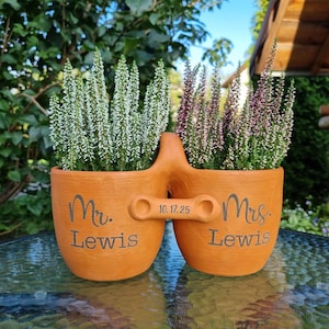 Peut inclure: Un pot en terre cuite avec deux récipients reliés par une poignée. Chaque pot porte l'inscription "Mr. Lewis" et "Mrs. Lewis". La date "10.17.25" est sur la poignée. Les pots contiennent des plantes à fleurs blanches et violettes.