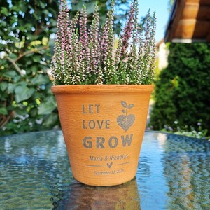Könnte beinhalten: Ein terrakottafarbener Blumentopf mit den Worten "LET LOVE GROW" und einem herzförmigen Pflanzendesign. Der Topf enthält eine blühende Pflanze mit rosa und grünem Laub. Die Namen "Maria & Nicholas" und das Datum "September 20, 2025" sind ebenfalls auf dem Topf aufgedruckt.