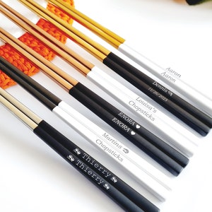 K&ouml;nnte beinhalten: Eine Sammlung personalisierter Essst&auml;bchen in verschiedenen Farben, darunter Gold, Schwarz und Silber. Jedes Essst&auml;bchen-Set verf&uuml;gt &uuml;ber eingravierte Namen und Daten. Die Essst&auml;bchen werden auf einer wei&szlig;en Oberfl&auml;che mit einem orangefarbenen Strickartikel im Hintergrund pr&auml;sentiert.