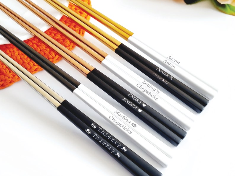 K&ouml;nnte beinhalten: Eine Sammlung personalisierter Essst&auml;bchen in verschiedenen Farben, darunter Gold, Schwarz und Silber. Jedes Essst&auml;bchen-Set verf&uuml;gt &uuml;ber eingravierte Namen und Daten. Die Essst&auml;bchen werden auf einer wei&szlig;en Oberfl&auml;che mit einem orangefarbenen Strickartikel im Hintergrund pr&auml;sentiert.