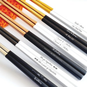 K&ouml;nnte beinhalten: Eine Auswahl an personalisierten Essst&auml;bchen in Schwarz, Wei&szlig; und Gold. Die Essst&auml;bchen sind mit Namen, Daten und S&auml;tzen wie "Sushi + Movie" und "We go together like a pair of chopsticks" graviert.