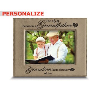 May include: A tan leather photo frame with a picture of a grandfather and grandson reading a book together. The frame has the text "The love between a Grandfather & Grandson lasts forever" with a heart symbol and the name "Liam" below.