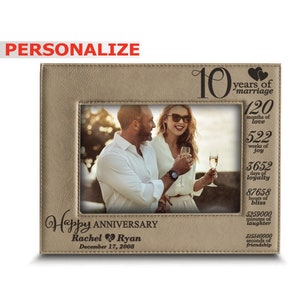 May include: A brown leather photo frame with a picture of a couple toasting with champagne. The frame has a gold inscription that reads "10 years of marriage" and lists the number of months, weeks, days, hours, minutes, and seconds that make up 10 years. The inscription also includes the text "Happy Anniversary Rachel Ryan December 17, 2008".