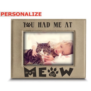 May include: A tan leather photo frame with a cat and a child in the photo. The frame has the text "You had me at Meow" with a paw print and the name "Lucy" in the bottom.