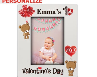Puede incluir: Marco de fotos blanco con el texto "Emma's 1st" y "Valentine's Day". El marco tiene una foto de una niña y está decorado con globos rojos en forma de corazón y una ilustración de oso de peluche. La palabra "PERSONALIZE" está en la parte superior.