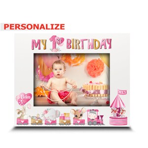 May include: A white picture frame with a pink and gold "My 1st Birthday" banner. The frame has a photo of a baby girl in a pink dress and a pearl necklace. The frame also has a pink train with animal characters and a pink and white carousel.