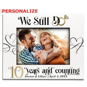 May include: A white photo frame with a gold and black design. The frame has a picture of a couple smiling and hugging. The text "We Still Do" is at the top of the frame. The text "10 years and counting" is at the bottom of the frame. The text "Darian & Lilian April 4, 20XX" is below the text "10 years and counting".