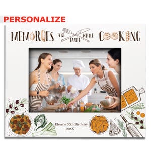 May include: White photo frame with a picture of four people cooking. The frame has a white background with a black border and the text "Memories are made while cooking" in black. The frame also has a black and white illustration of a fork and knife crossed over each other.