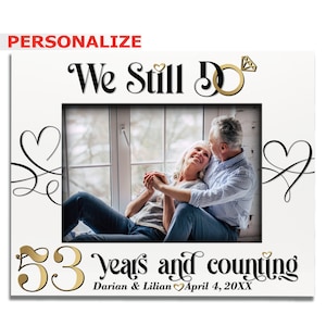 May include: A white photo frame with a gold ring and diamond design. The text "We Still Do" is written in black cursive font. The frame has a photo of an older couple sitting together. The text "53 years and counting" is written in black cursive font with a gold number 53. The text "Darian & Lilian April 4, 20XX" is written in black cursive font.