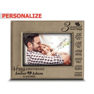 May include: A tan leather photo frame with a picture of a couple inside. The frame has a personalized message that says "Happy Anniversary Amber & Adam 11.05.20XX". The frame also has a decorative inscription that says "3 years of marriage" and lists the number of months, weeks, days, hours, minutes, and seconds that have passed in their marriage.