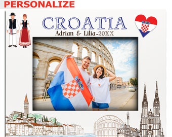 Può includere: Una cornice per foto bianca con un disegno di un paesaggio urbano croato, che include una chiesa, una torre e un anfiteatro. La cornice ha un cuore rosso con la bandiera croata al suo interno e il testo "CROATIA Adrian & Lilia-20XX" nella parte superiore. Ci sono due figure di cartoni animati di persone con abiti tradizionali croati sul lato sinistro della cornice.