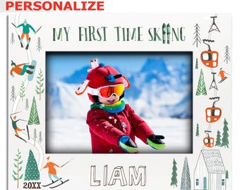 Puede incluir: Un marco de fotos blanco con un borde de escena de invierno. El marco tiene una foto de un niño con una chaqueta roja y gafas protectoras. El texto "My First Time Skiing" está en la parte superior del marco y el nombre "Liam" está en la parte inferior.